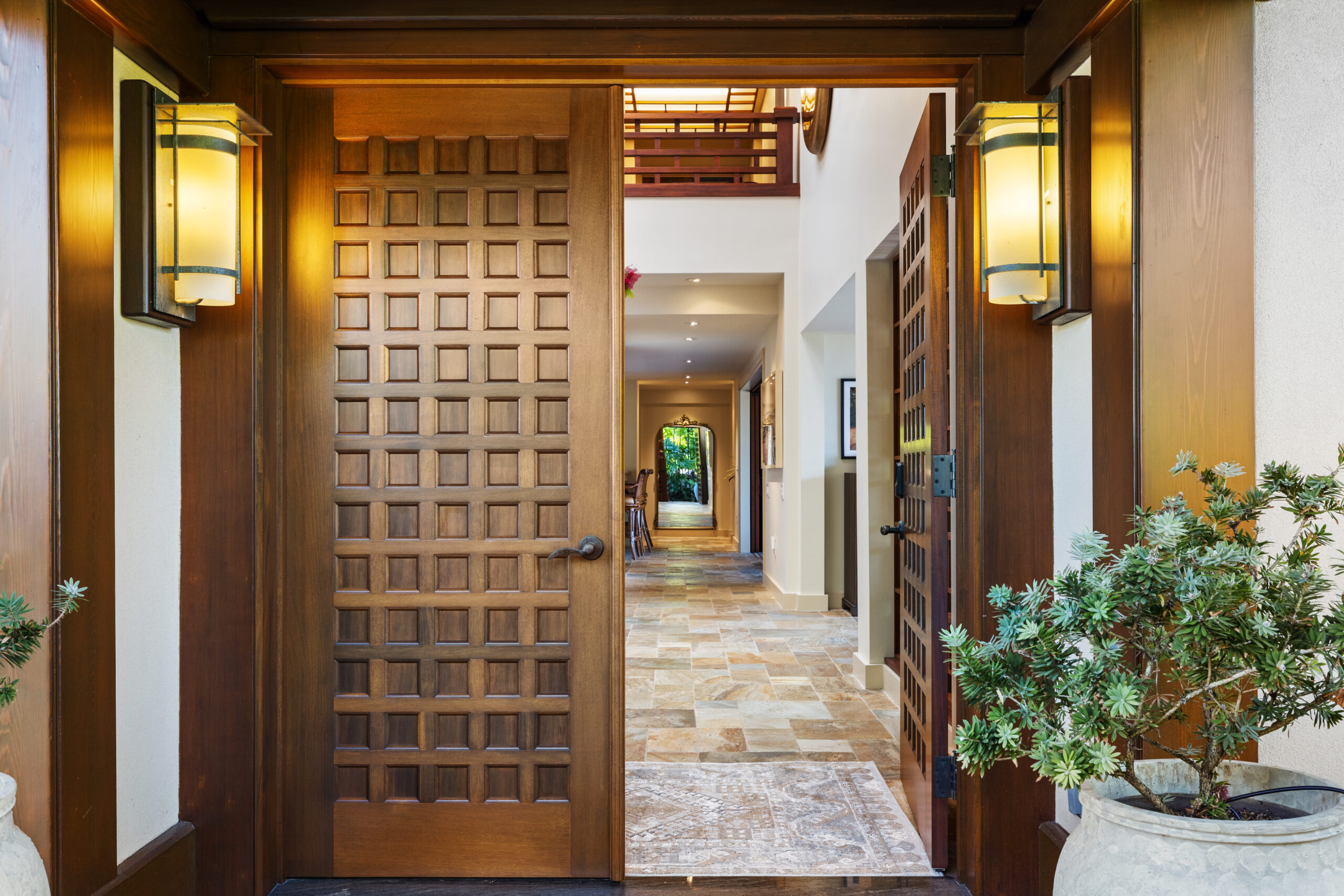 Elegant villa entrance hallway featuring rich wood beam ceiling, geometric wood screen panels, matching lantern-style wall sconces, natural stone tile flooring, white walls with dark wood trim, and glimpses of tropical garden views through the corridor leading to bright outdoor spaces.