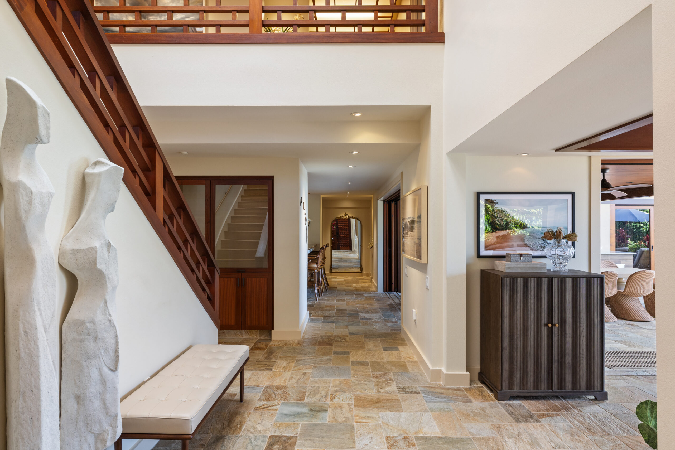 Elegant two-story foyer featuring dramatic geometric wood staircase with matching railings, natural stone tile flooring, skylight above, modern bench seating, sculptural art piece, and wood louvered doors with tropical orchids.