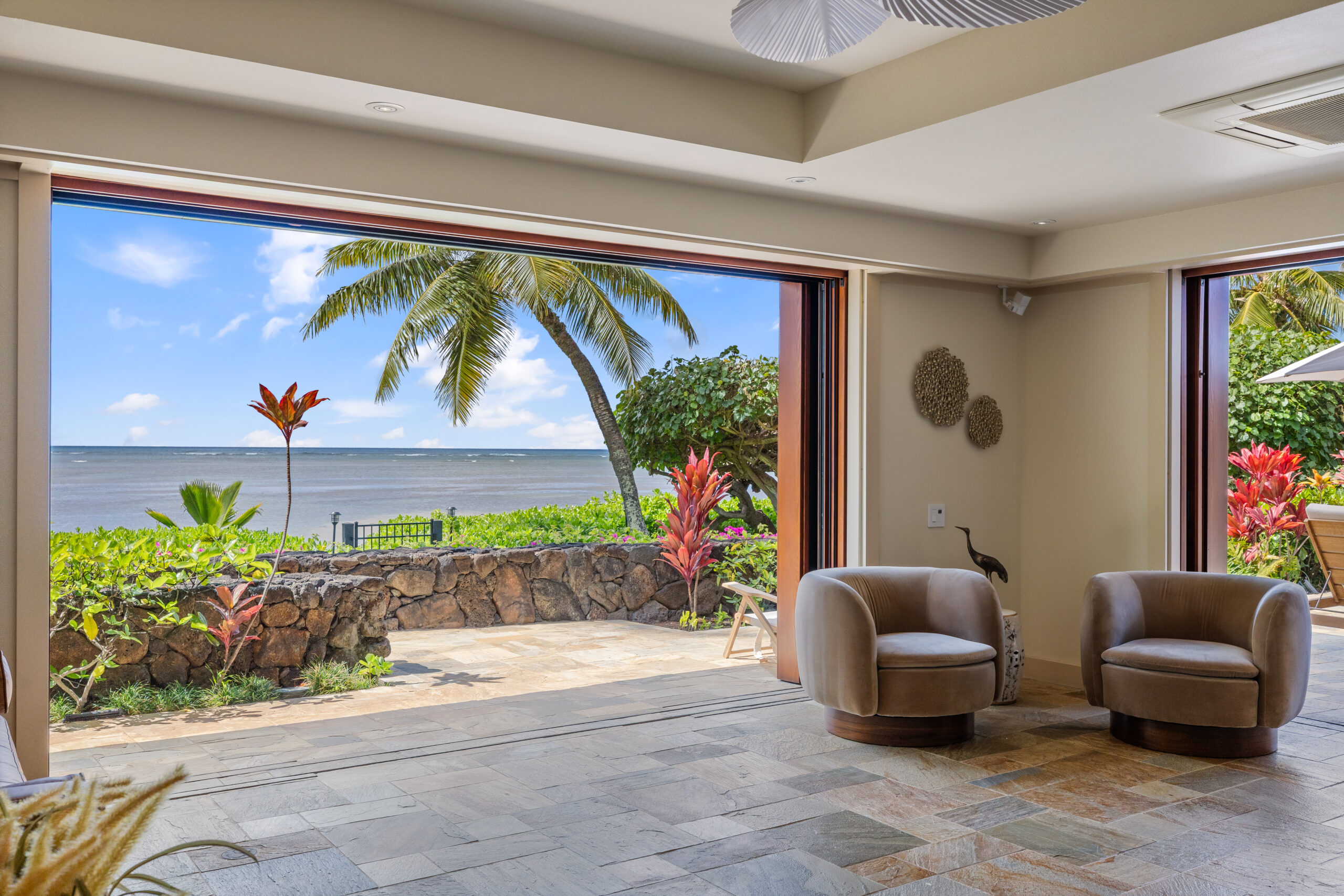 Spacious living area featuring natural stone tile flooring, wooden louvered closet doors, floor-to-ceiling windows with tropical garden views, modern round swivel chairs, built-in seating, and white ceiling fan.