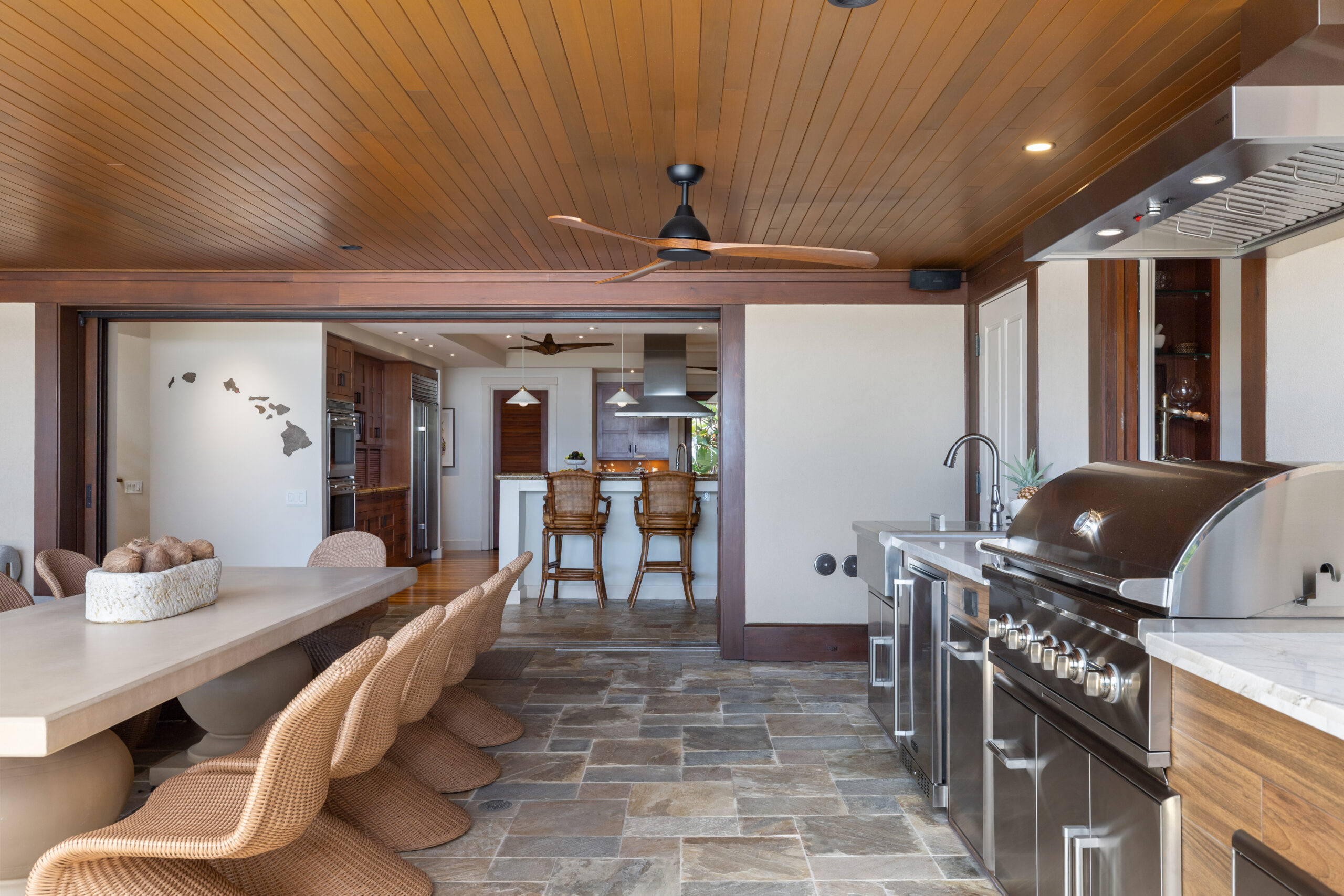 Luxury covered outdoor dining area with wood plank ceiling, ceiling fans, and wicker dining chairs around large table, featuring full outdoor kitchen with stainless steel appliances, stone tile flooring, and panoramic ocean views framed by tropical landscaping including red ti plants, palm trees, and infinity pool edge.