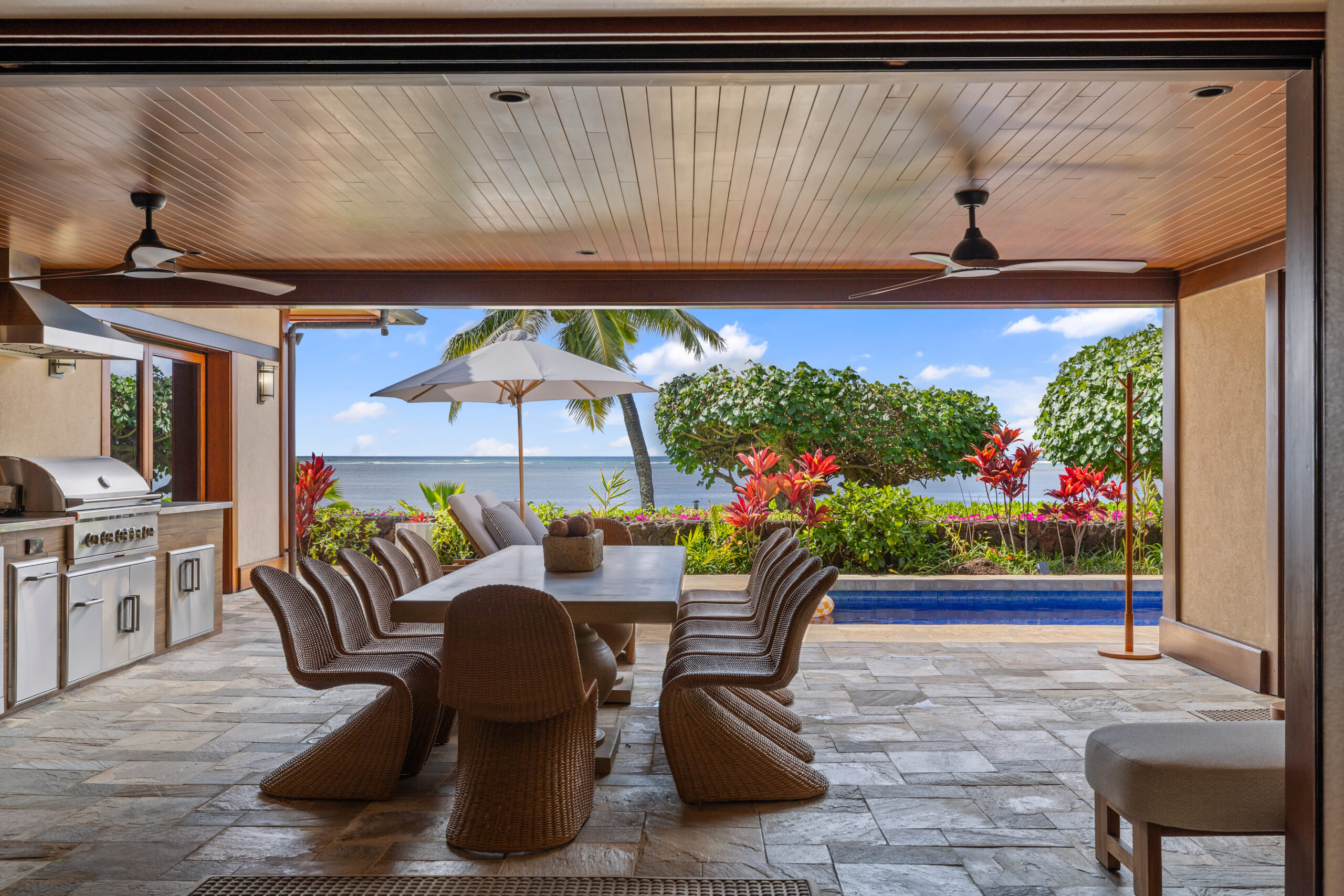 Luxury covered outdoor dining area with wood plank ceiling, ceiling fans, and wicker dining chairs around large table, featuring full outdoor kitchen with stainless steel appliances, stone tile flooring, and panoramic ocean views framed by tropical landscaping including red ti plants, palm trees, and infinity pool edge.