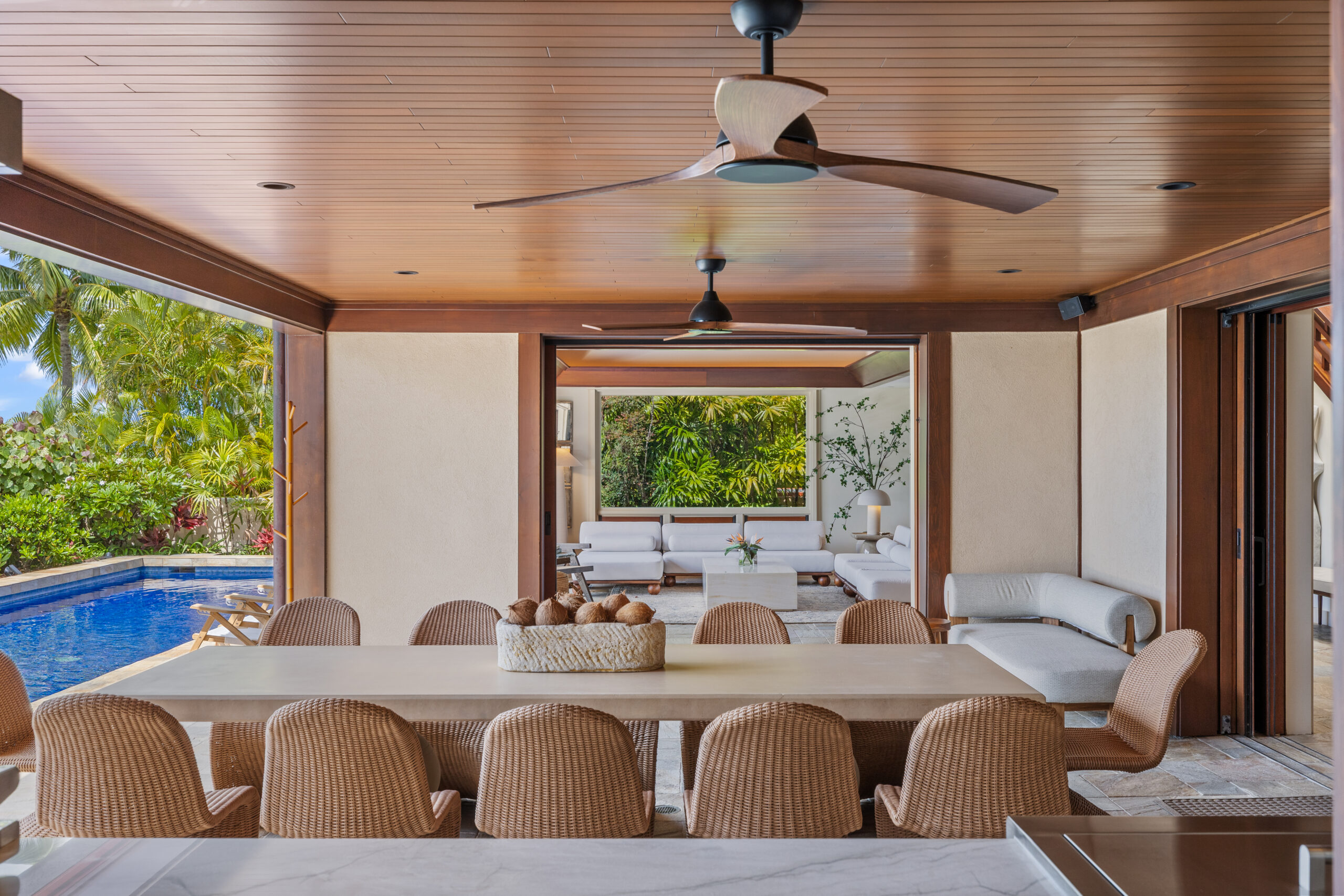 Luxury covered outdoor dining area with wood plank ceiling, ceiling fans, and wicker dining chairs around large table, featuring full outdoor kitchen with stainless steel appliances, stone tile flooring, and panoramic ocean views framed by tropical landscaping including red ti plants, palm trees, and infinity pool edge.