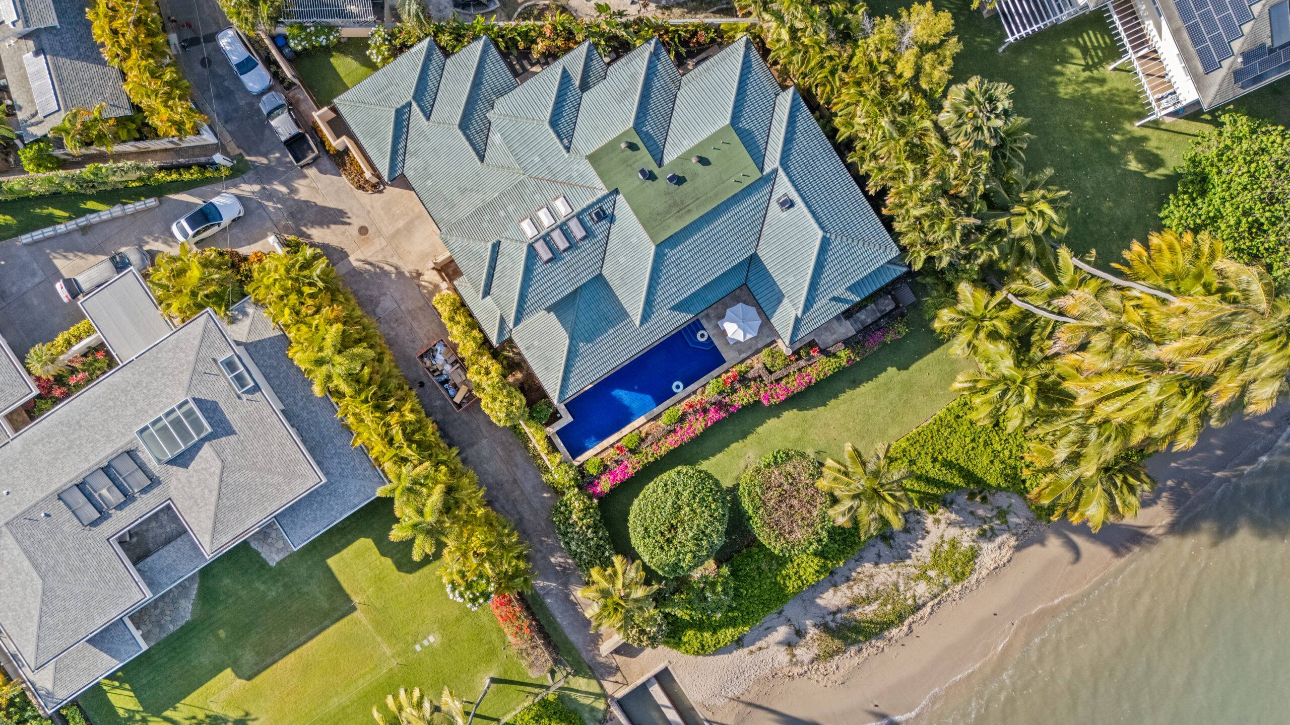 Elevated view of luxury villa nestled among lush tropical hillside community with palm trees, manicured landscapes, neighboring homes, and dramatic cloudy sky backdrop across rolling green terrain.