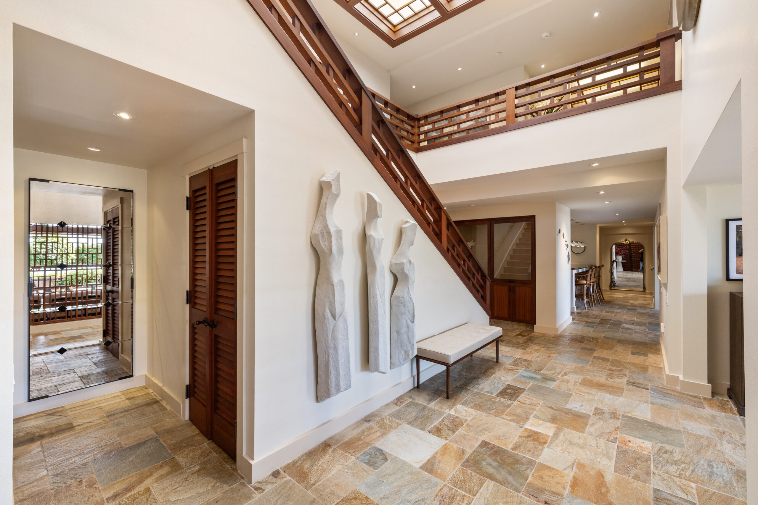 Elegant two-story foyer featuring dramatic geometric wood staircase with matching railings, natural stone tile flooring, skylight above, modern bench seating, sculptural art piece, and wood louvered doors with tropical orchids.