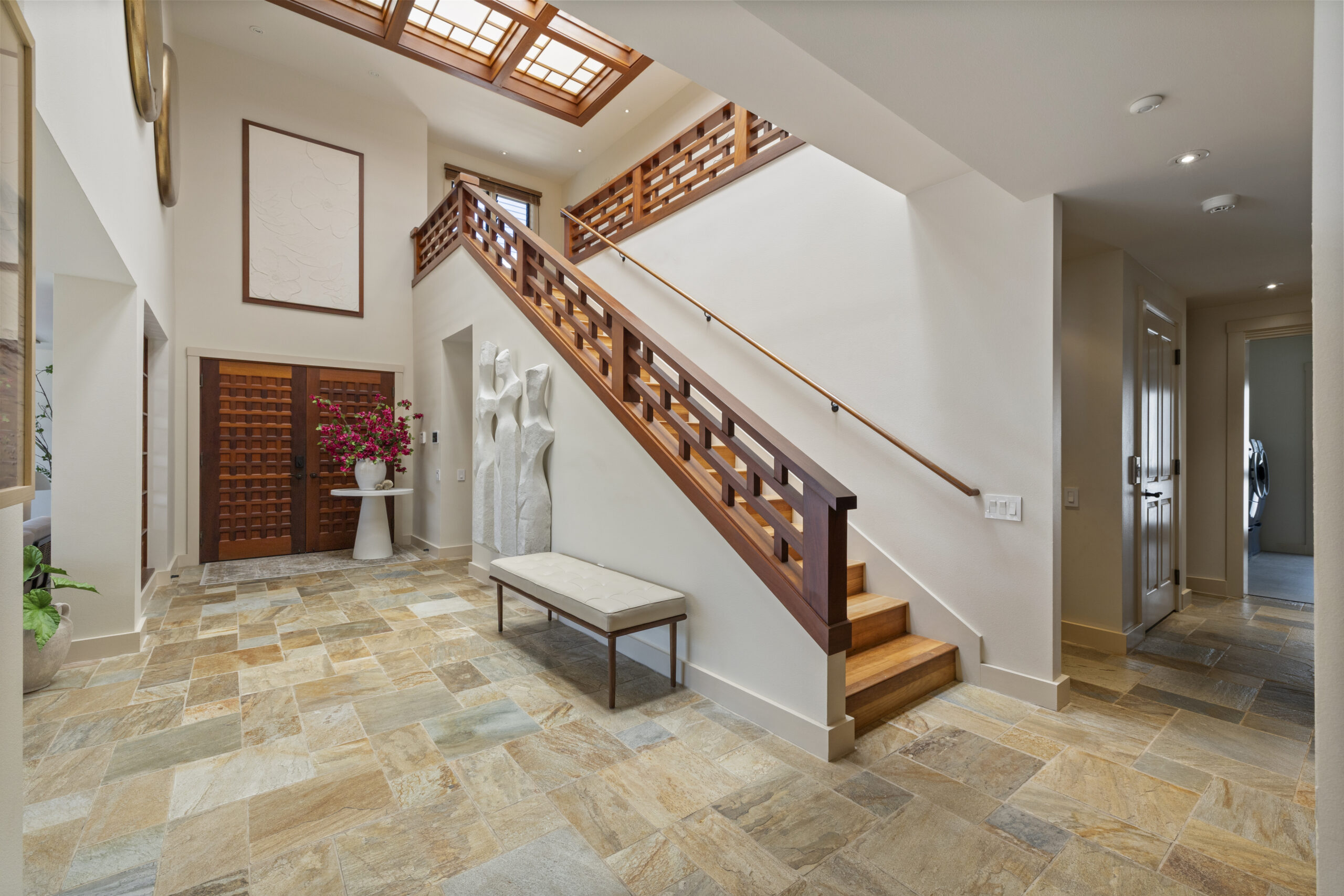 Elegant two-story foyer featuring dramatic geometric wood staircase with matching railings, natural stone tile flooring, skylight above, modern bench seating, sculptural art piece, and wood louvered doors with tropical orchids.