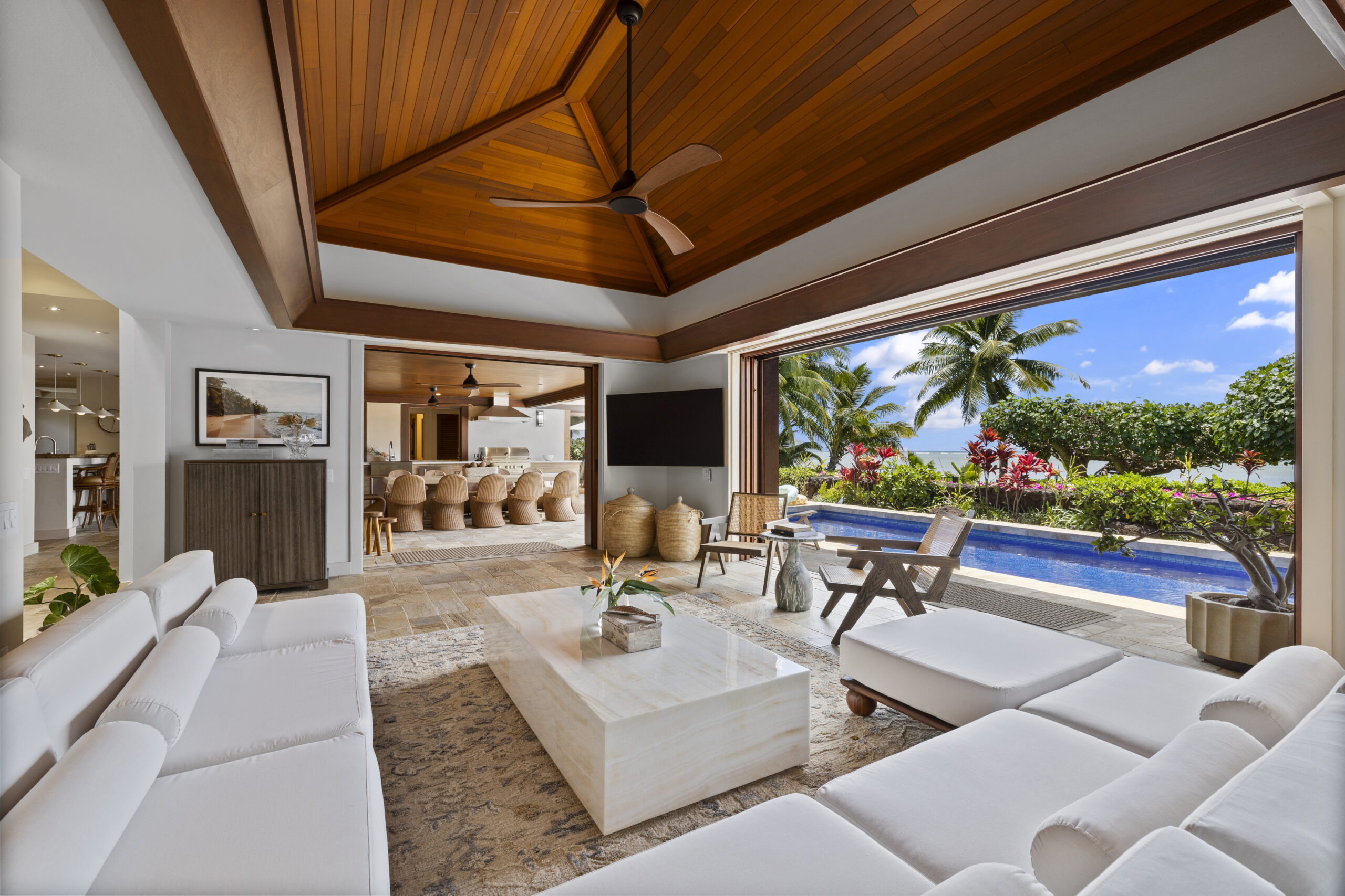 Luxurious open-concept living space featuring dramatic vaulted wood ceiling with exposed beams, white sectional seating, geometric wood screen divider, natural stone flooring, and seamless indoor-outdoor flow to dining area.