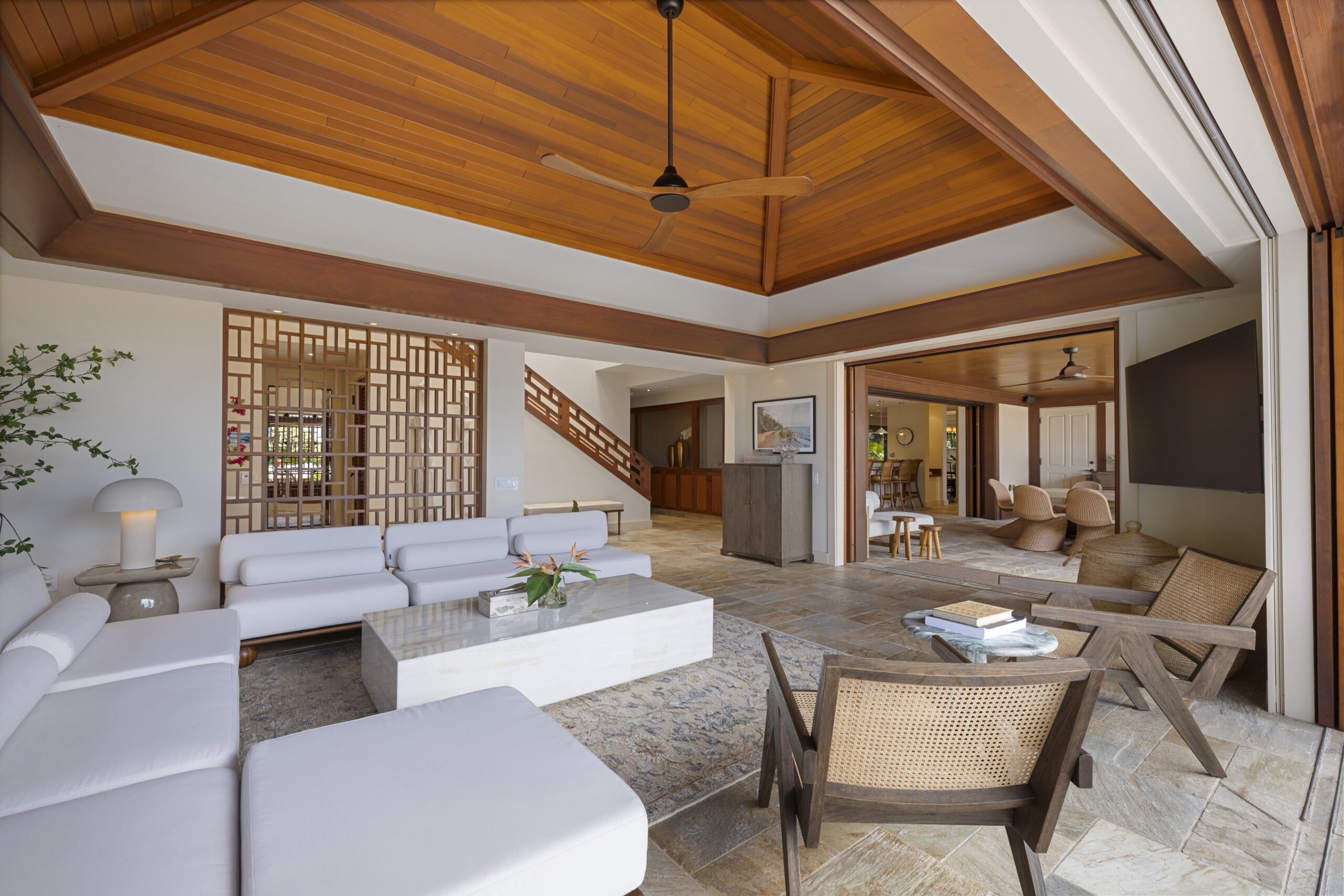 Luxurious open-concept living space featuring dramatic vaulted wood ceiling with exposed beams, white sectional seating, geometric wood screen divider, natural stone flooring, and seamless indoor-outdoor flow to dining area.
