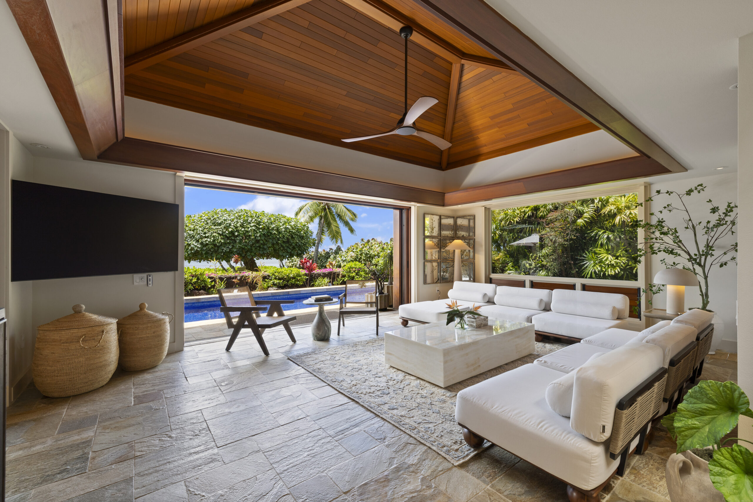 Luxurious open-concept living space featuring dramatic vaulted wood ceiling with exposed beams, white sectional seating, geometric wood screen divider, natural stone flooring, and seamless indoor-outdoor flow to dining area.