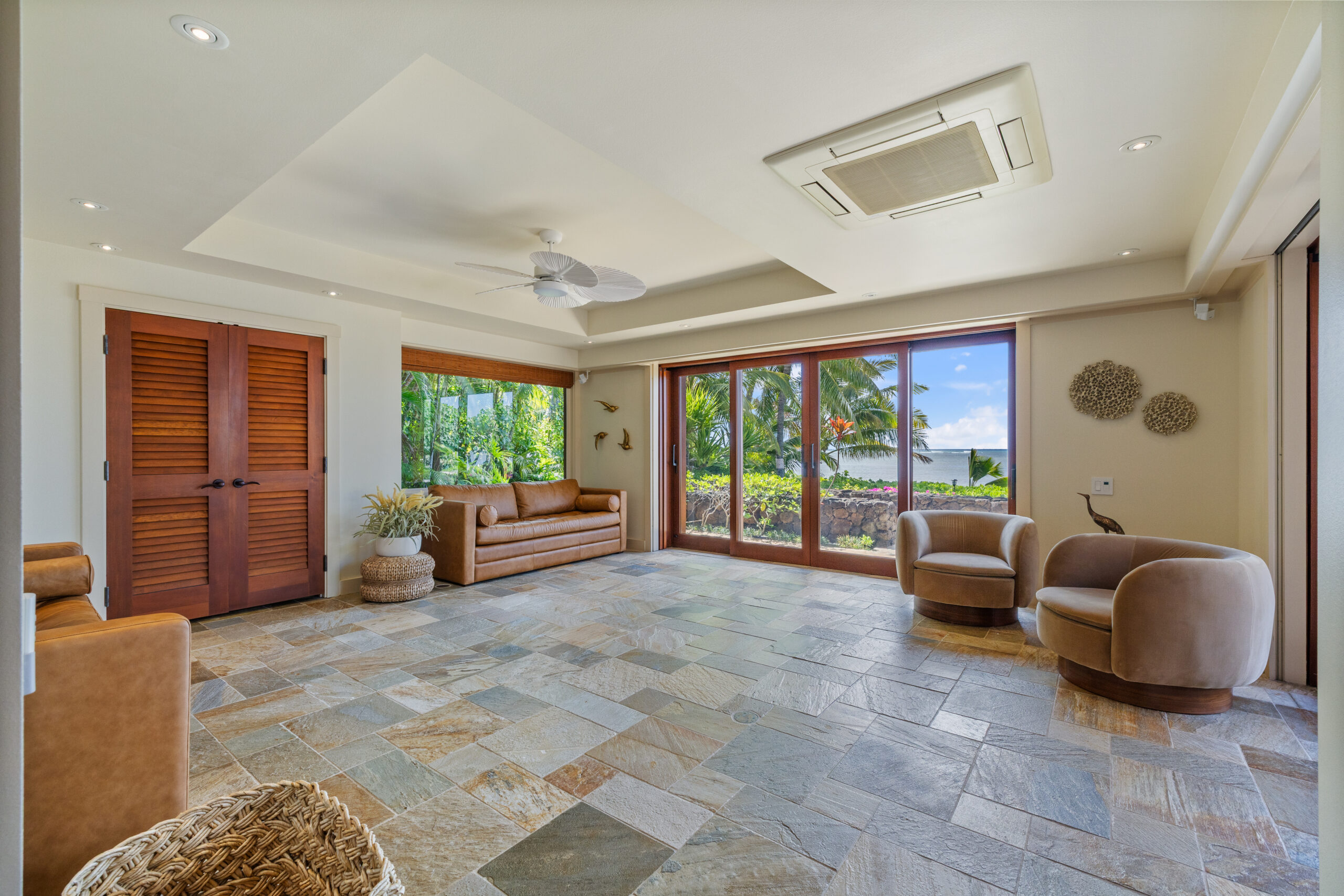Spacious living area featuring natural stone tile flooring, wooden louvered closet doors, floor-to-ceiling windows with tropical garden views, modern round swivel chairs, built-in seating, and white ceiling fan.