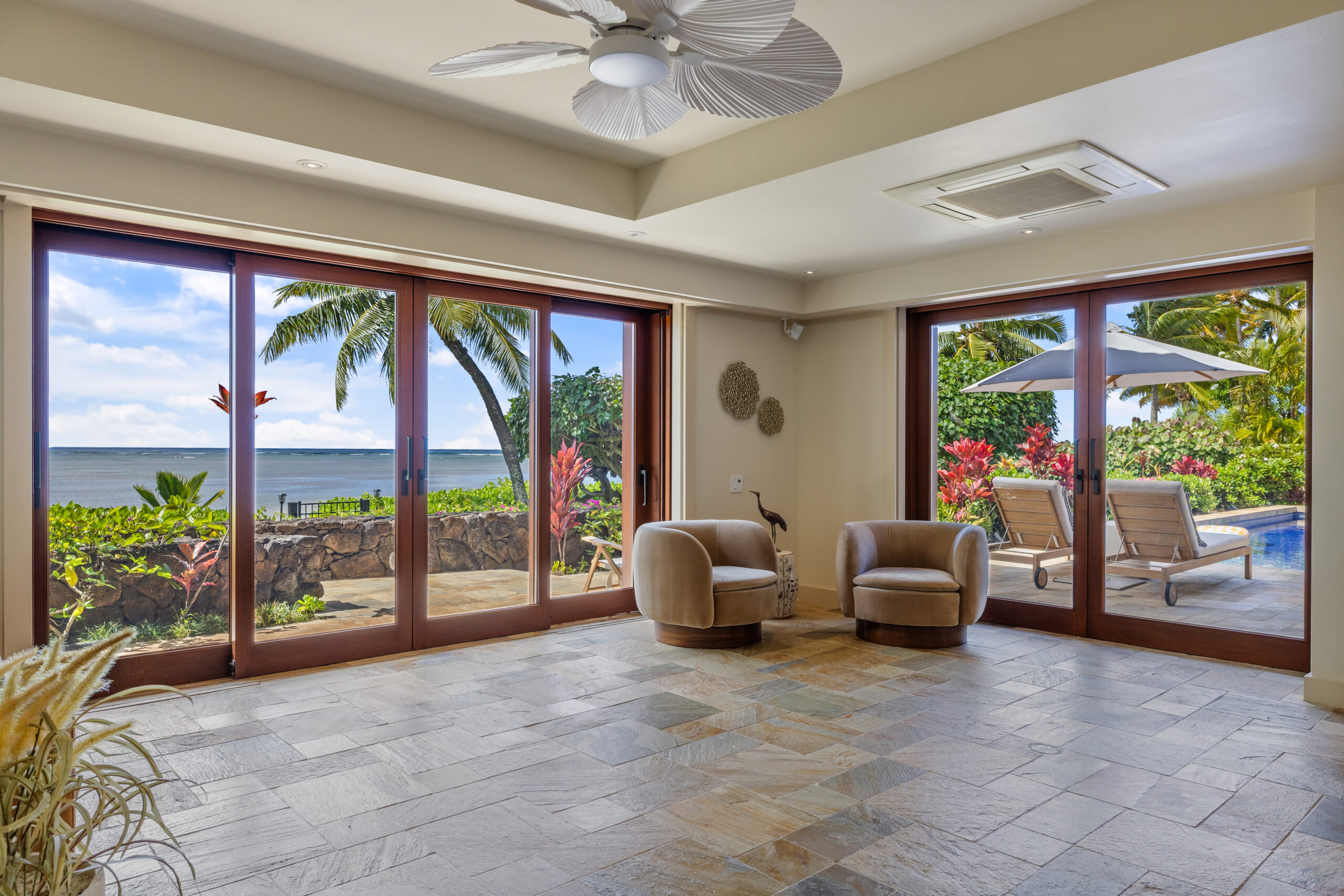 Spacious living area featuring natural stone tile flooring, wooden louvered closet doors, floor-to-ceiling windows with tropical garden views, modern round swivel chairs, built-in seating, and white ceiling fan.