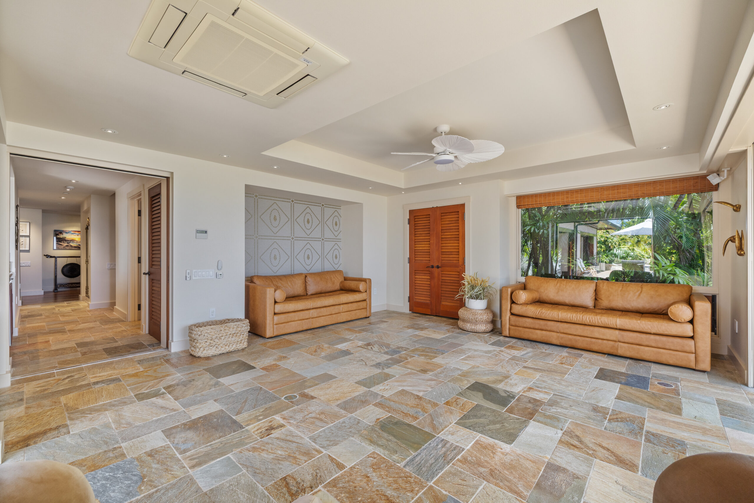 Spacious living area featuring natural stone tile flooring, wooden louvered closet doors, floor-to-ceiling windows with tropical garden views, modern round swivel chairs, built-in seating, and white ceiling fan.