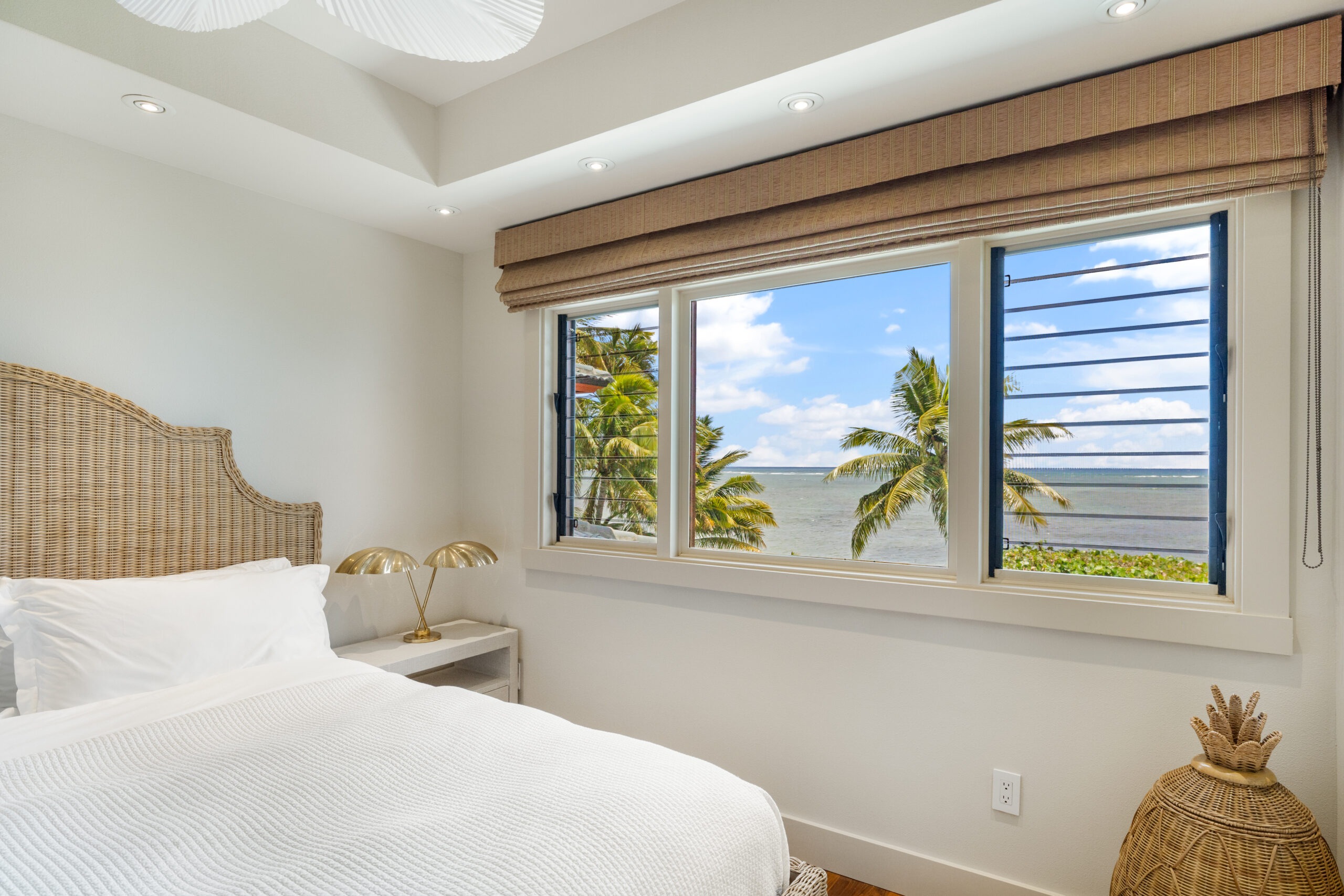 Serene king bedroom featuring natural woven rattan headboard, white linens, bedside table with tropical plant, and louvered windows framing palm trees and blue sky views.