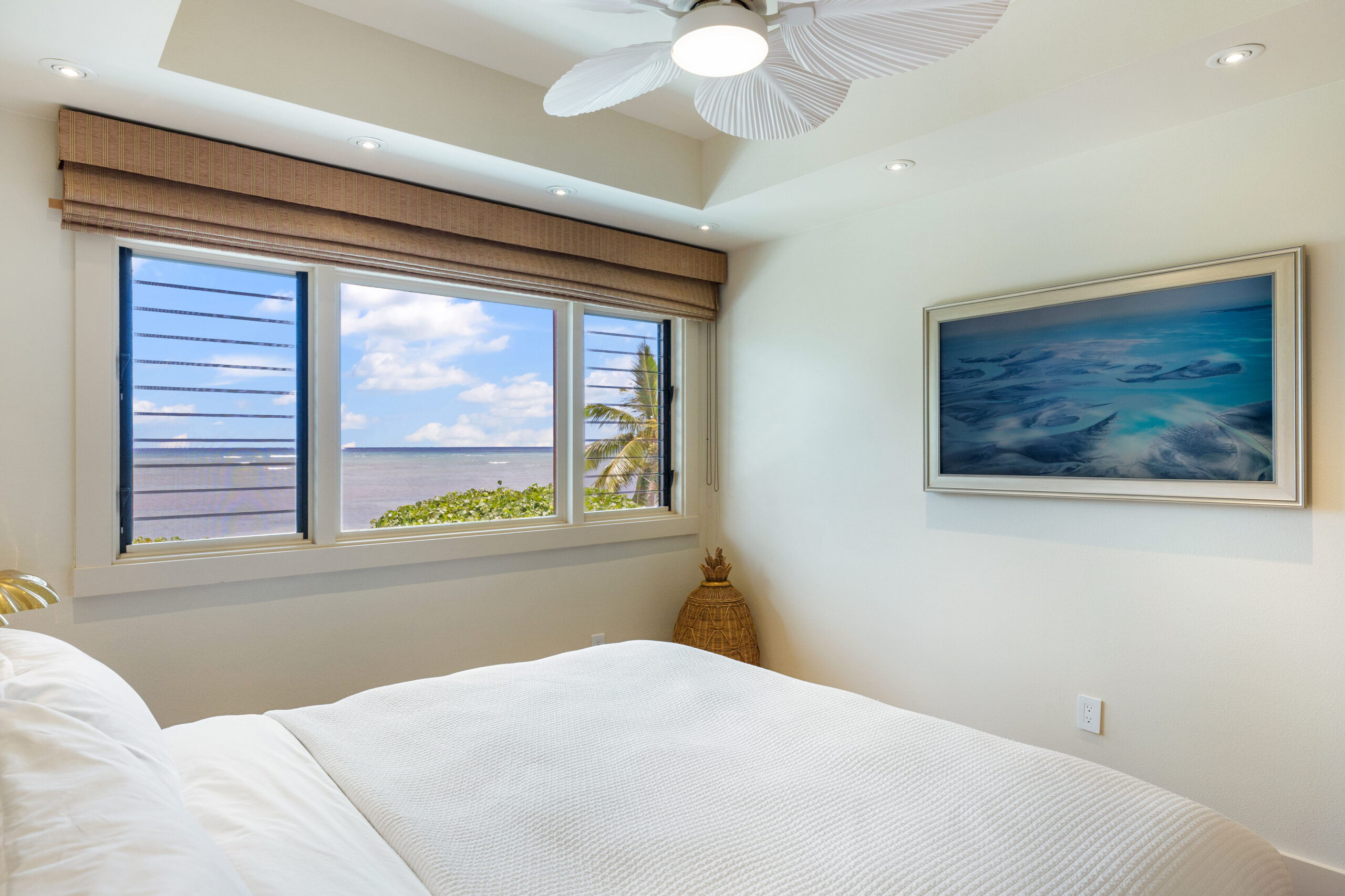 Bright king bedroom with white linens, louvered windows showcasing ocean and tropical landscape views, white ceiling fan, and framed ocean artwork on wall with clean modern design.