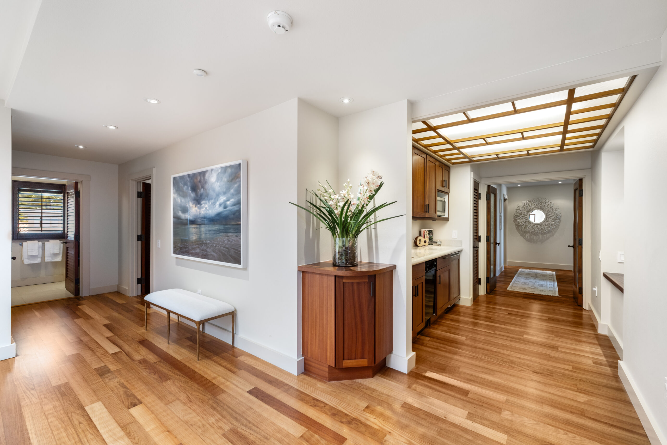 Luxurious guest wing hallway featuring rich hardwood floors, white walls with recessed lighting, custom wood cabinetry kitchenette with built-in appliances, dramatic geometric skylight with warm wood frame, modern white bench seating, ocean-themed artwork, fresh orchid arrangement on wood credenza, and view to guest bathroom with wooden shutters.