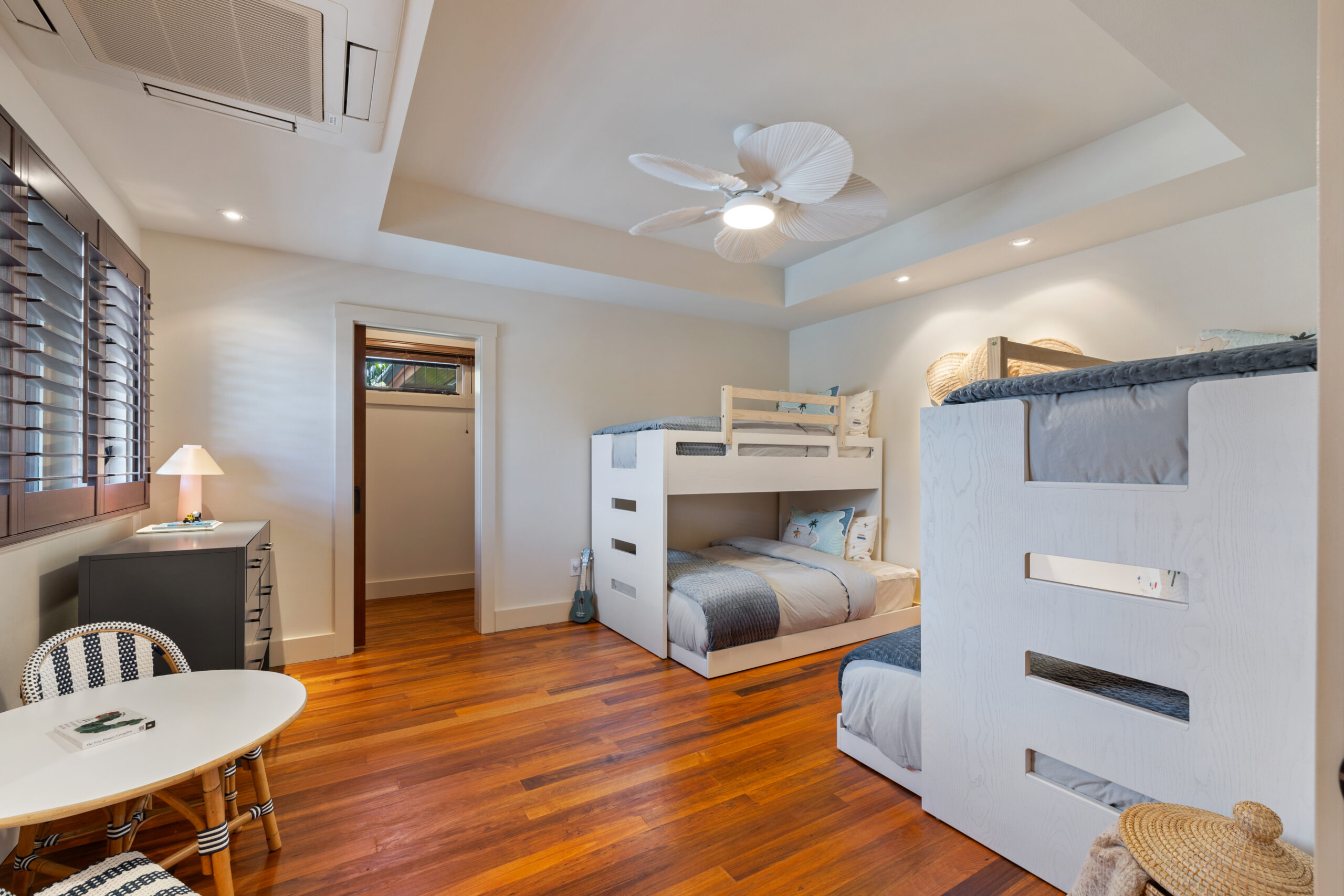 Bright bunk room with white built-in beds featuring full-size bottom bunk and twin-size top bunk, hardwood floors, white ceiling fan, round table with chairs, and louvered windows overlooking tropical gardens.