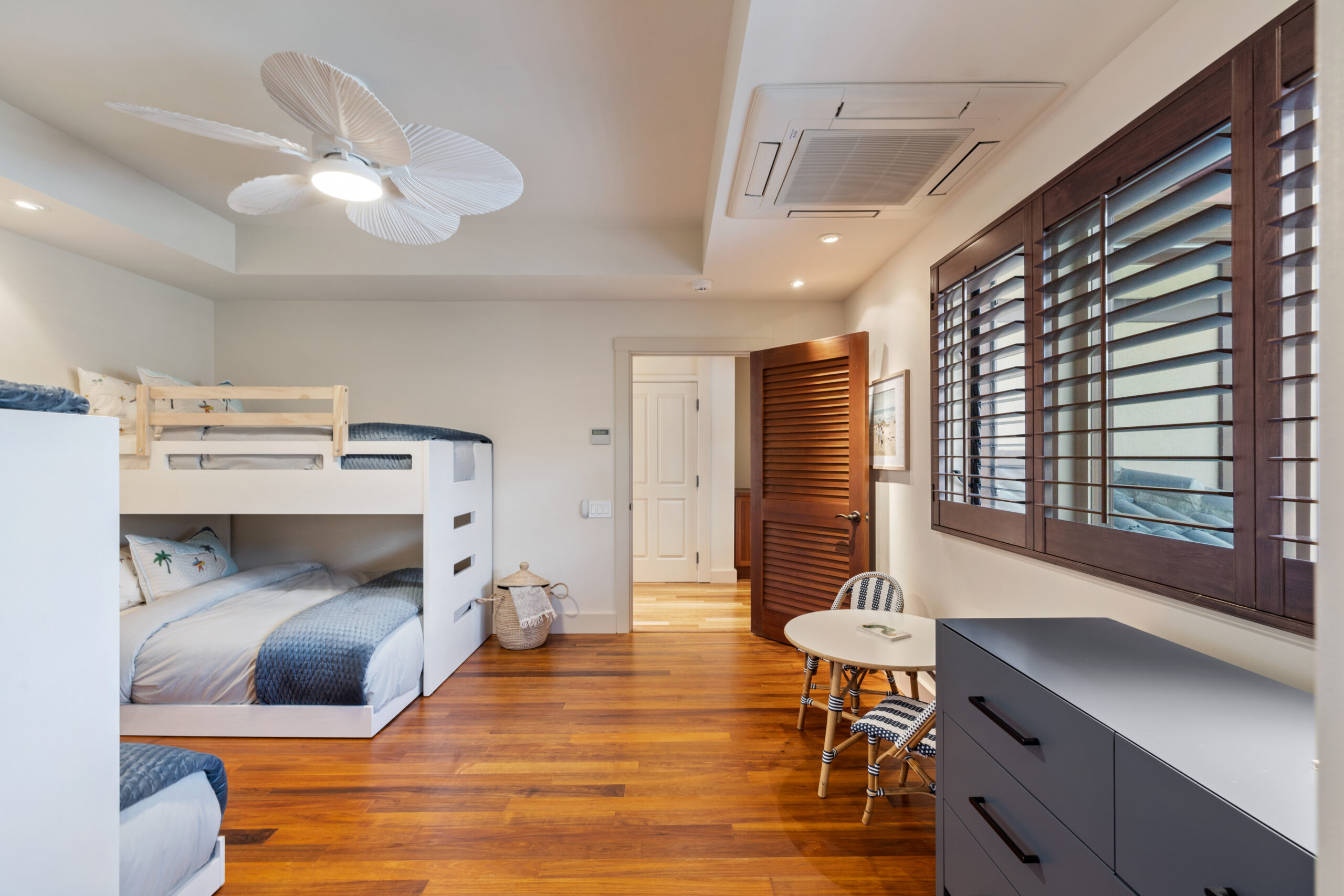 Modern bunk room featuring white built-in beds with full bottom bunk and twin top bunk, hardwood floors, dresser, round table with white chair, white ceiling fan, and louvered windows with garden views.