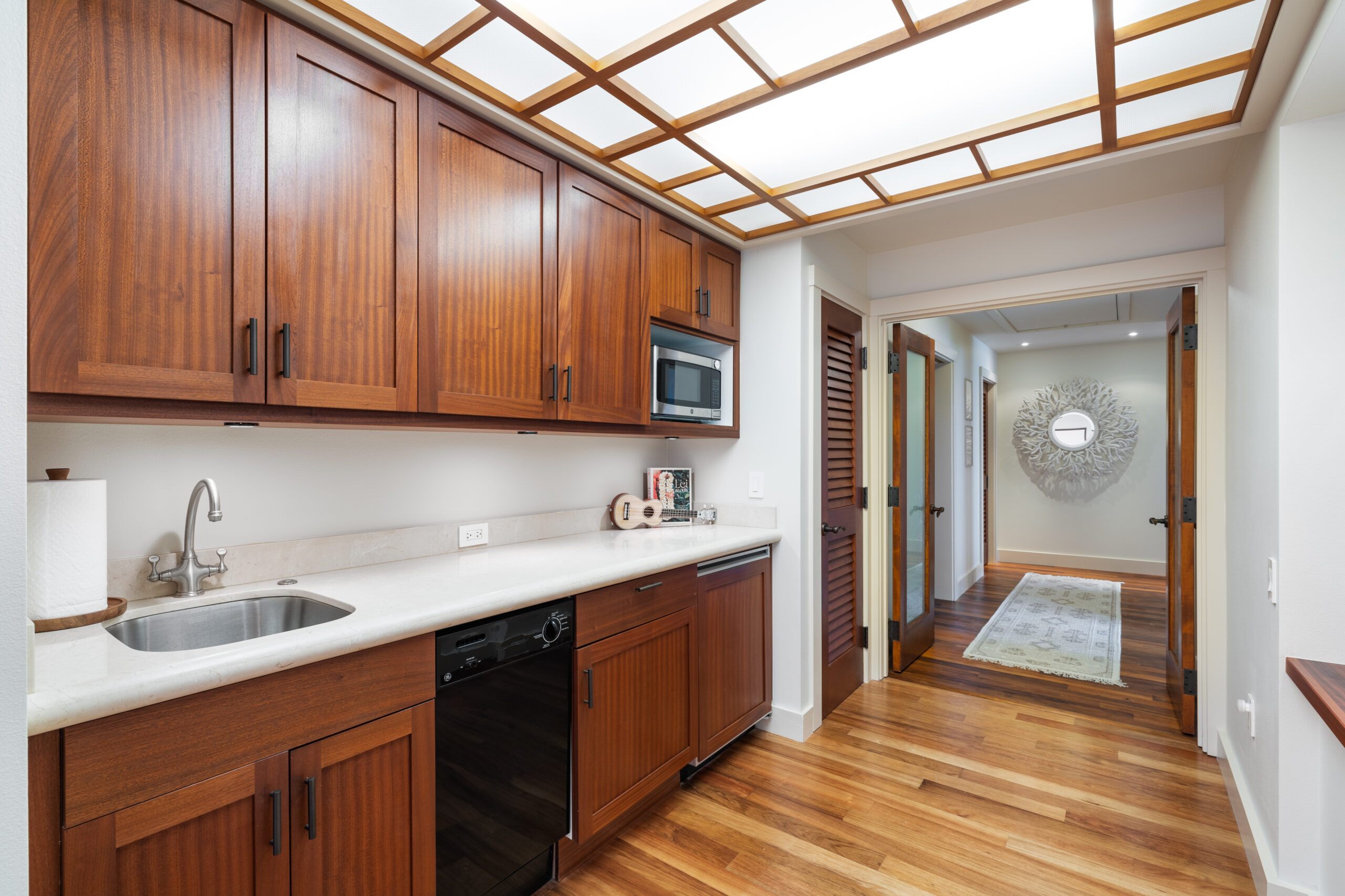 Elegant guest kitchen featuring rich wood cabinetry with white quartz countertops, stainless steel sink and appliances, stunning coffered skylight ceiling with natural lighting, warm hardwood flooring, white walls, and open connection to hallway with decorative mirror and louvered closet doors.