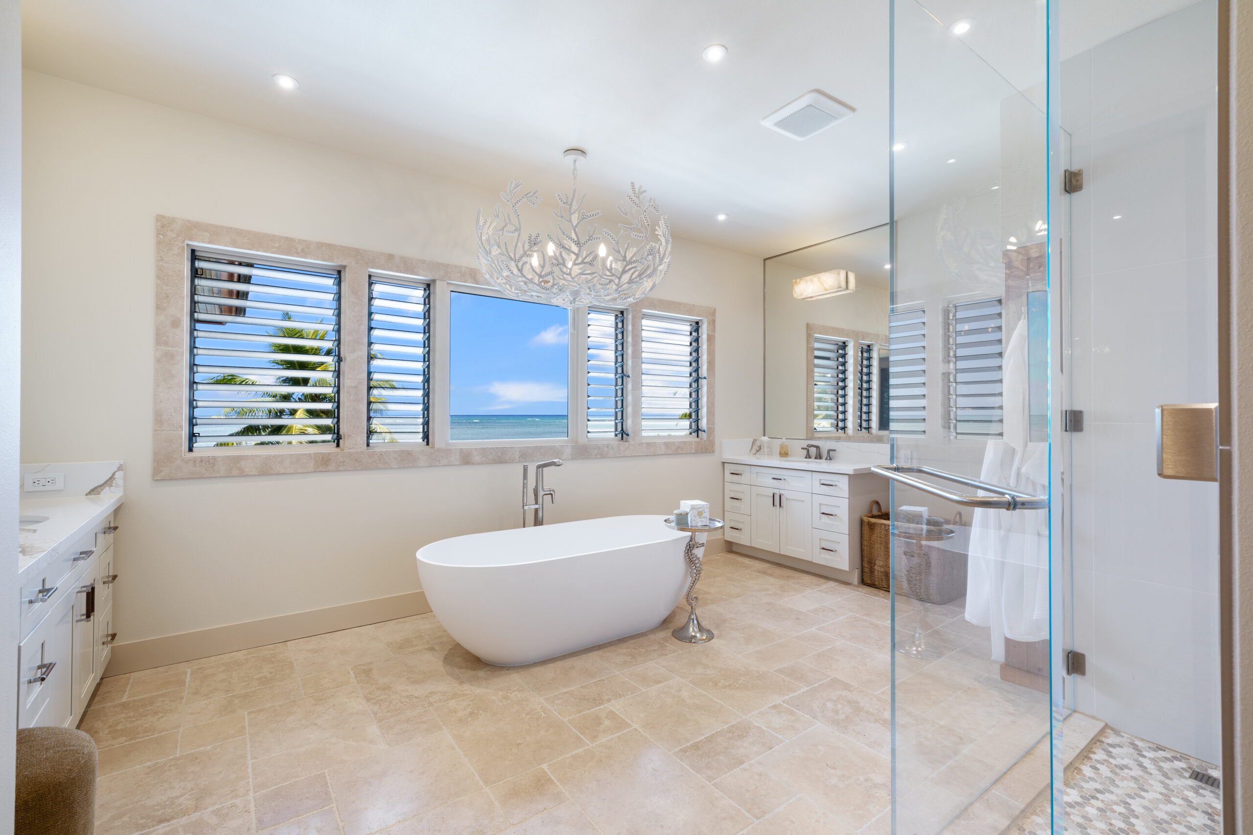 Luxurious spa bathroom featuring freestanding white soaker tub positioned beneath louvered windows with direct ocean views, coral branch chandelier, dual vanity, and glass-enclosed shower.