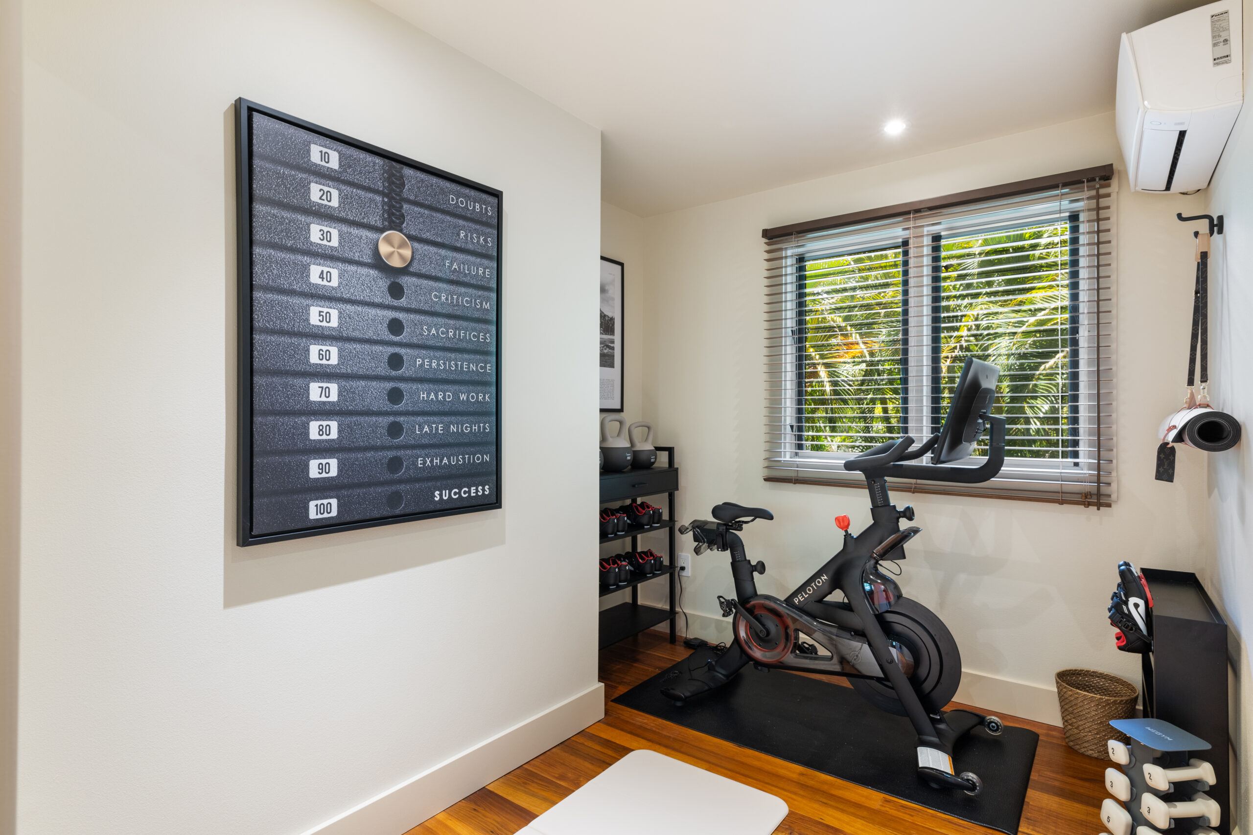 Dedicated fitness room featuring Peloton Bike+ on rubber flooring, wall-mounted workout guide chart, weight storage rack, and tropical garden views through louvered windows with hardwood floors.