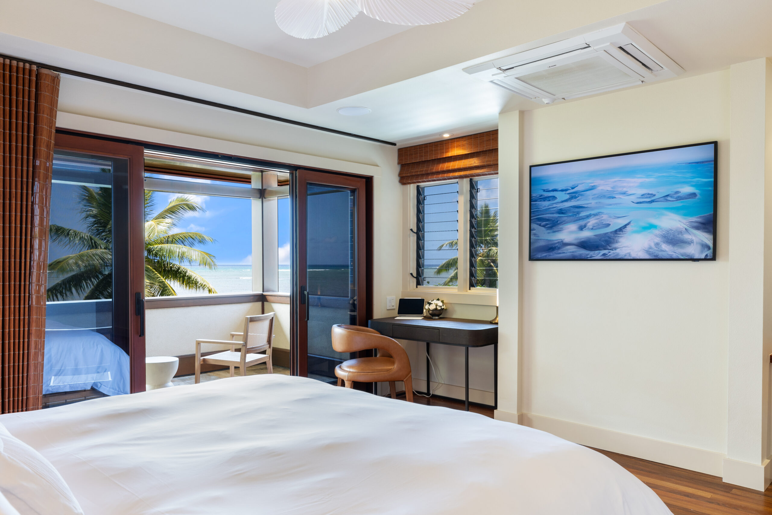 Luxurious bedroom with king bed, built-in work desk, wall-mounted TV displaying ocean scenes, and direct access to private balcony with outdoor seating and palm tree views through floor-to-ceiling windows.