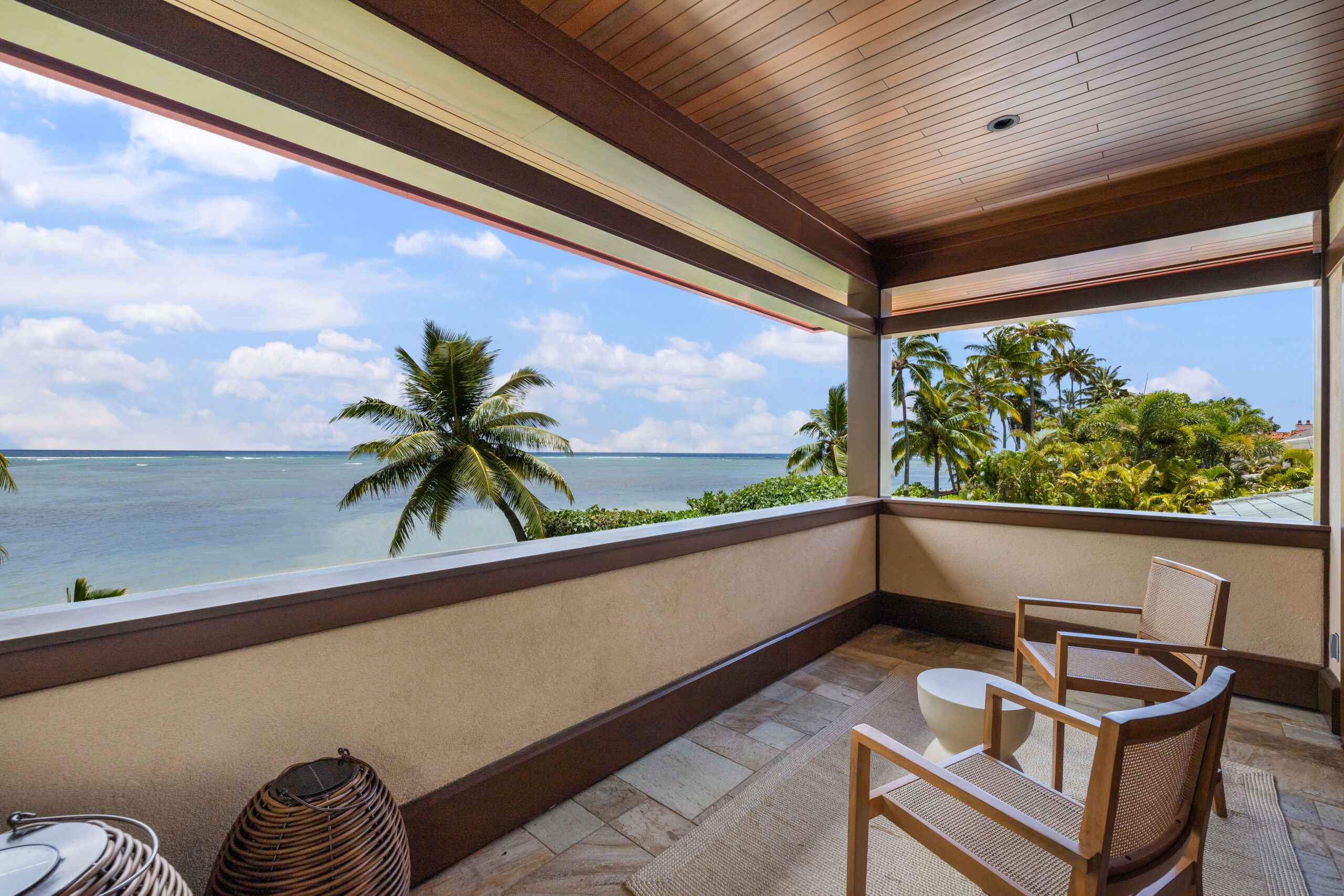 Covered oceanfront balcony with wood beam ceiling, stone tile flooring, comfortable seating for two, and unobstructed panoramic views of turquoise waters, palm trees, and tropical coastline.