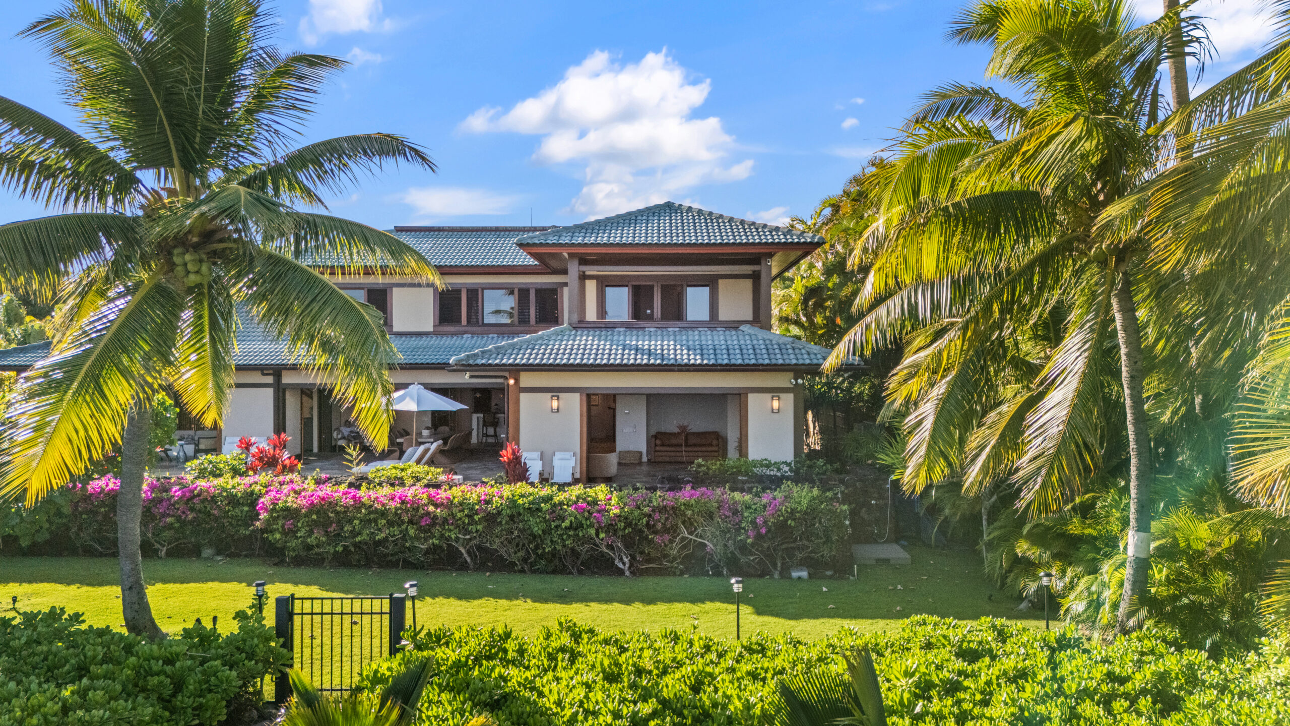 Elegant oceanfront backyard featuring manicured lawn, mature shade trees with rope hammock, natural lava rock borders, well-maintained tropical hedging, landscape lighting, and direct beach access with pristine sand and turquoise ocean waters under blue sky with white clouds.
