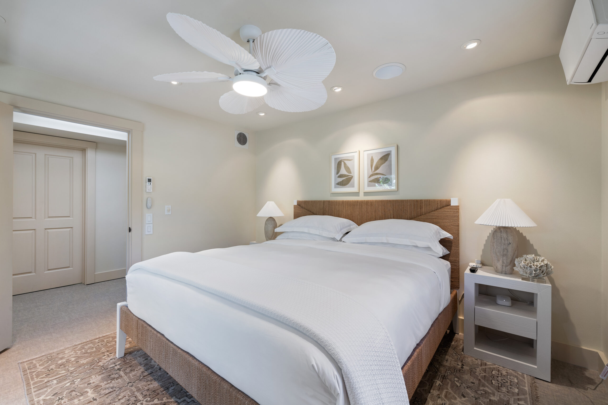 Clean and bright guest bedroom featuring king bed with white linens, wooden headboard, white ceiling fan, bedside tables with lamps, coastal artwork, and white bifold closet doors.
