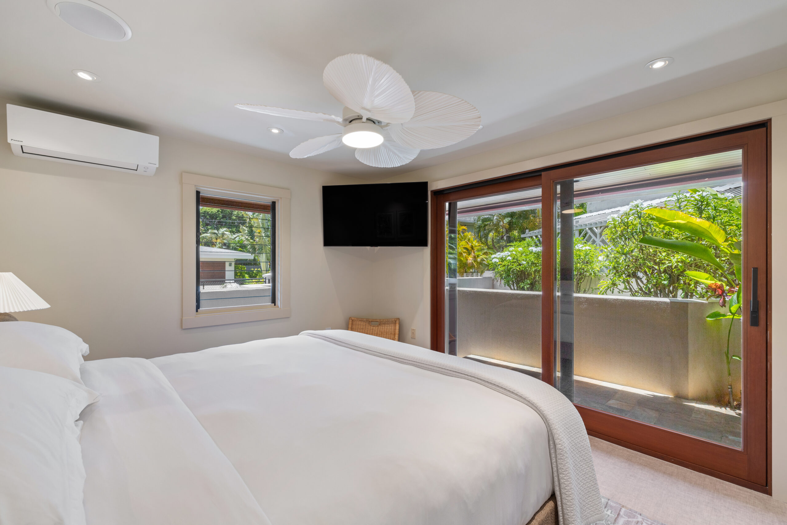 Serene king bedroom on lower level featuring white linens, wall-mounted TV, white ceiling fan, sliding glass doors opening to private garden terrace, and lush tropical landscaping views.