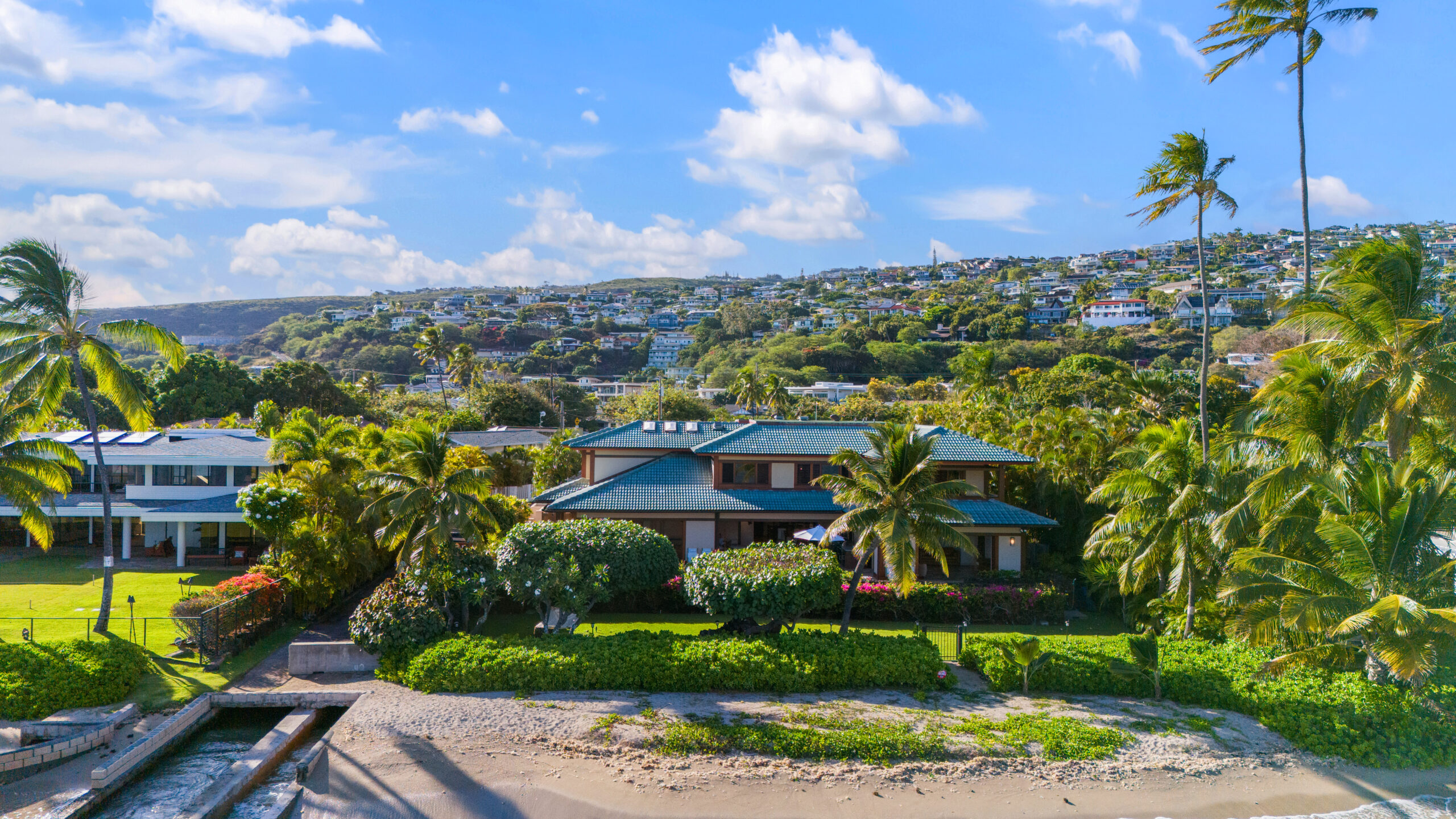 Elevated view of luxury villa nestled among lush tropical hillside community with palm trees, manicured landscapes, neighboring homes, and dramatic cloudy sky backdrop across rolling green terrain.