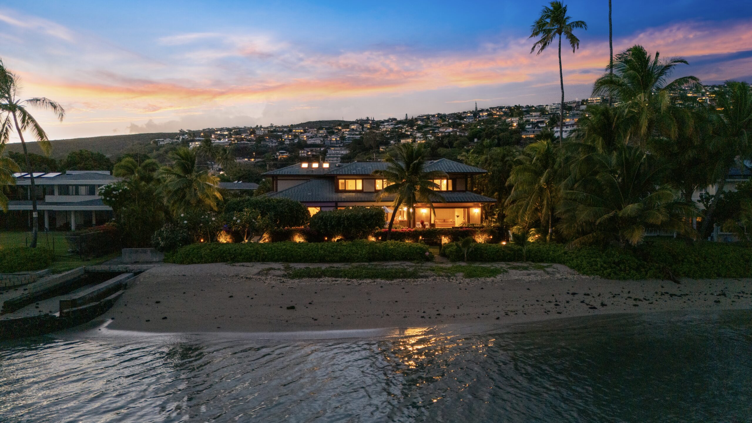 Elevated view of luxury villa nestled among lush tropical hillside community with palm trees, manicured landscapes, neighboring homes, and dramatic cloudy sky backdrop across rolling green terrain.