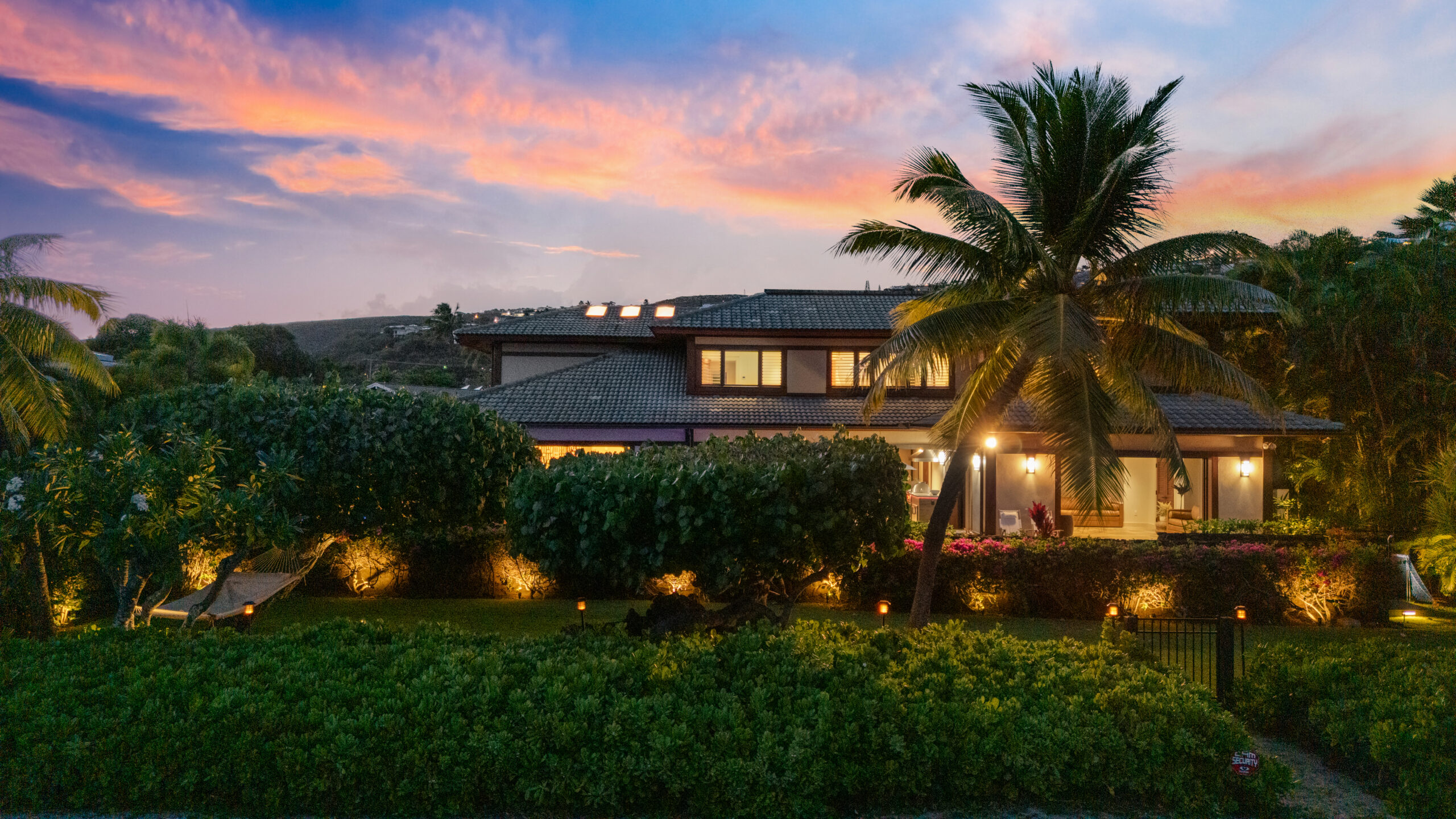 Stunning two-story contemporary villa at twilight featuring dark tile roofing, warm interior lighting glowing through large windows, surrounded by mature coconut palms and lush tropical landscaping with dramatic uplighting illuminating trees and gardens, set against vibrant sunset sky with pink, orange and purple clouds.
