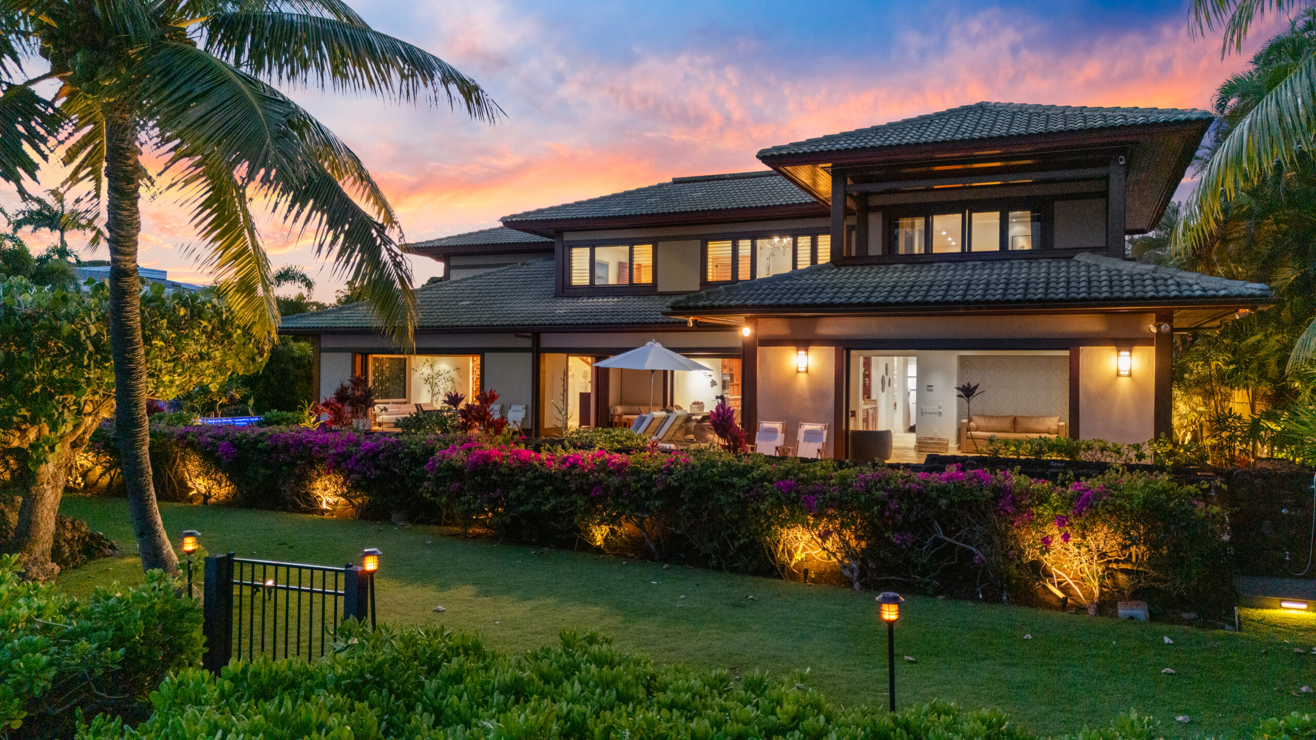 Two-story modern tropical villa with expansive glass windows, warm interior lighting, surrounded by palm trees and manicured tropical landscaping with dramatic sunset sky in pink and orange hues.