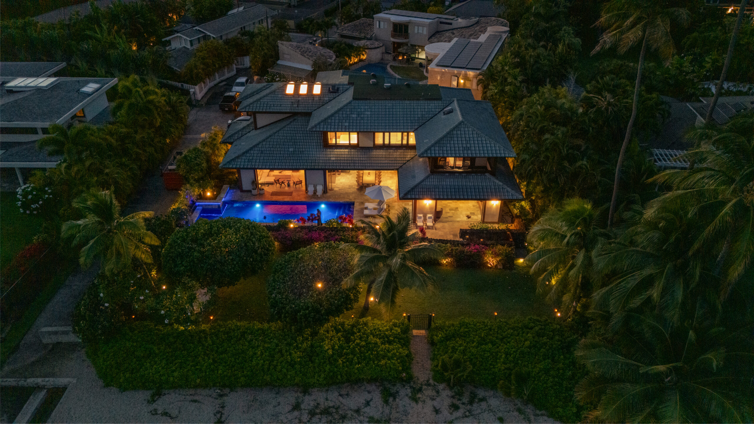 Elevated view of luxury villa nestled among lush tropical hillside community with palm trees, manicured landscapes, neighboring homes, and dramatic cloudy sky backdrop across rolling green terrain.